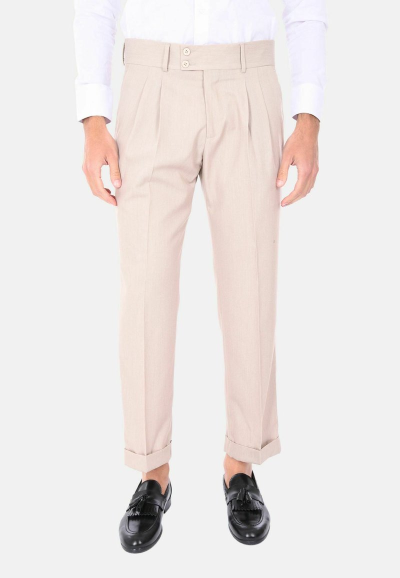 Pantaloni beige su misura con doppia piega frontale, chiusura a due bottoni, orli arrotolati e una superficie liscia, abbinati a mocassini neri.