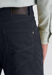 Dunkelblaues Cordhosen aus strukturiertem Material, mit einer Gesäßtasche, einem Leder-Logo-Patch und einem schlanken Schnitt. Das Hemd hat ein hellgrünes Muster.