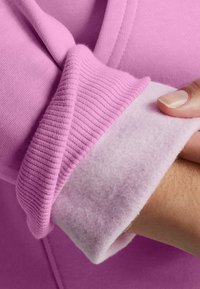 Una mano che tiene il polsino di un indumento in pile rosa, mostrando il morbido tessuto interno e il bordo esterno a coste.