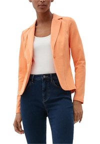 Blazer naranja claro con un solo botón, solapas con muesca y dos bolsillos laterales. Llevado sobre una camiseta de tirantes blanca y combinado con jeans oscuros.