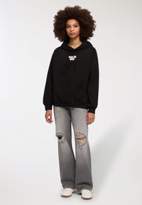 Felpa nera con cappuccio e la scritta "BACIAMI ADESSO" stampata sul davanti, abbinata a jeans grigi a gamba larga strappati e sneakers bianche.