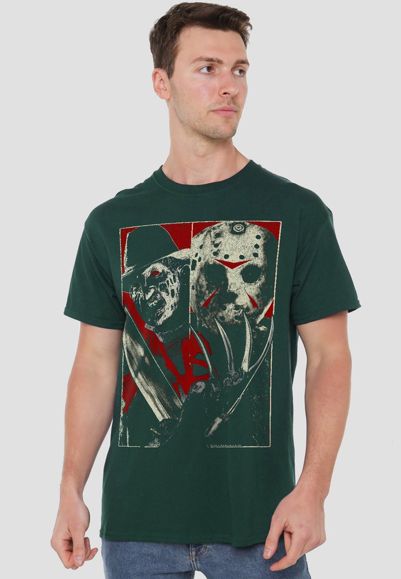 Man wearing dark green T-shirt with graphic of two masked horror characters holding weapons, standing against plain light background.