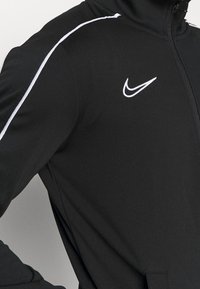 Black athletic jacket made of smooth fabric, featuring white accent stripes along the shoulders and a Nike logo on the chest.