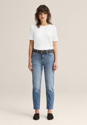 Kvinde med krøllet hår iført en helt hvid kortærmet skjorte, blå jeans med brunt bælte og mørke loafers, stående foran en neutral baggrund.