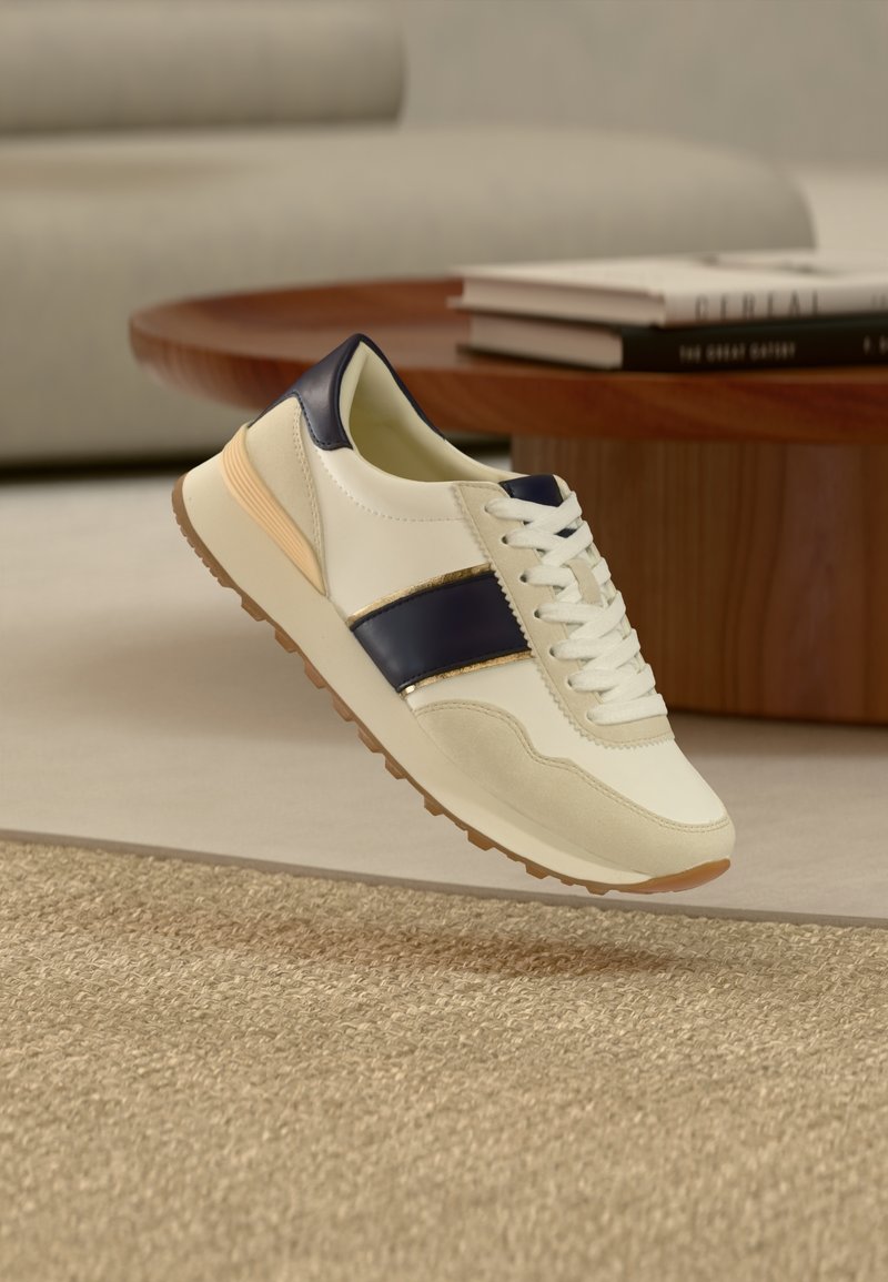 Sneaker bianca e blu navy con dettagli beige che fluttua sopra un tappeto intrecciato, accanto a un tavolino in legno con libri impilati.