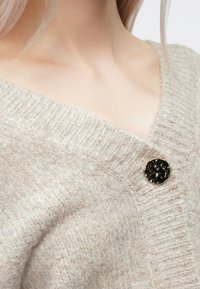 Világos bézs kötött kardigán V-nyakkal, fekete, texturált felületű, kerek gombbal és finom kontrasztos részletekkel.