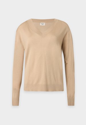 Vrouw met lang donker haar draagt een beige V-hals trui, wijde zwarte broek, bordeauxrode schoenen, houdt een opgevouwen bruine jas vast tegen een witte achtergrond.