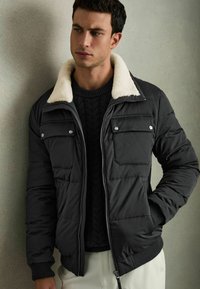 Jachetă puffer neagră cu guler din blană artificială albă, având două buzunare pe piept, închidere frontală cu fermoar și manșete cu dantelă. Purta o bluză închisă la culoare.