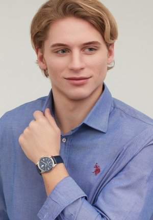 Young man in blue shirt looks to side, showing wristwatch with dark face and leather strap on raised arm.