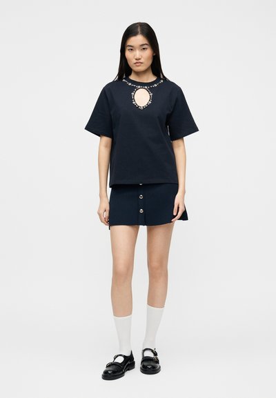 Γυναίκα με μαύρο τοπ με λαιμόκοψη διακοσμημένη με πολύτιμους λίθους και κοντή φούστα με κουμπιά, συνδυασμένη με λευκές κάλτσες και μαύρα παπούτσια.