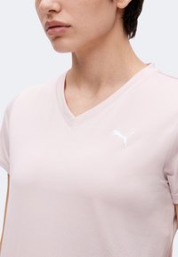 Γυναίκα που φοράει ροζ ανοιχτό T-shirt με V λαιμόκοψη και λευκό λογότυπο Puma στο αριστερό στήθος, μπροστά σε ένα απλό ανοιχτό φόντο.