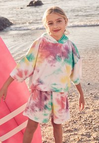 Tie-dye hættetrøje i pastelfarver (pink, gul, grøn) med korte ærmer og matchende shorts, lavet af blødt materiale, strandbaggrund.