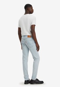 Helles blaugraues Denimjeans im Slim Fit, mit einem Lederetikett am Bund und Gesäßtaschen. Kombiniert mit einem weißen T-Shirt und schwarzen Schuhen.