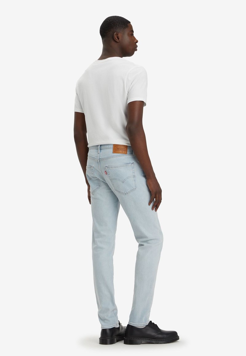 Jeans de mezclilla de color azul claro con un corte slim, que cuentan con una etiqueta de cuero en la cinturilla y bolsillos traseros. Combinados con una camiseta blanca y zapatos negros.