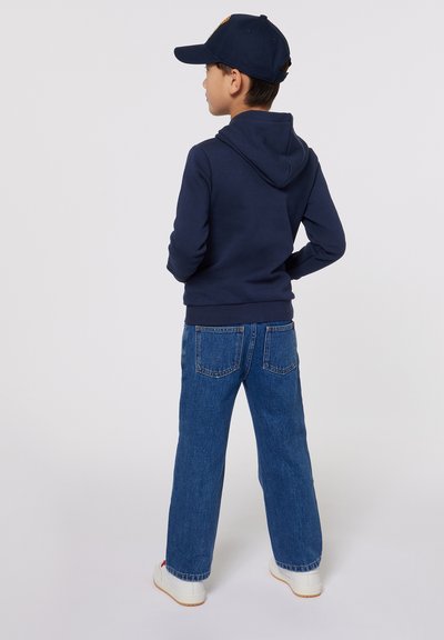 Hoodie marine avec une poche kangourou et une capuche, associé à un jean droit bleu clair. Baskets blanches avec des accents rouges.