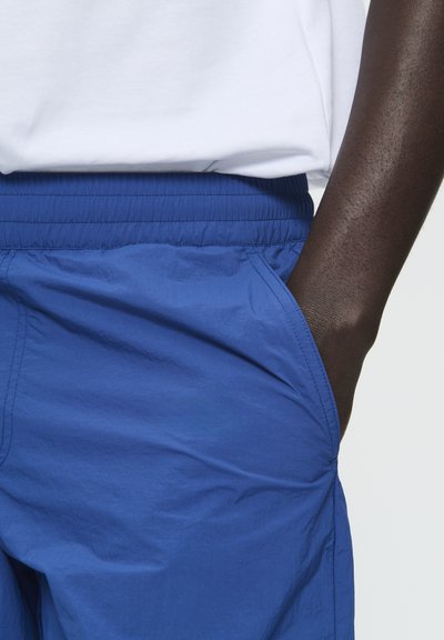 Blaue Shorts mit Gummibund und seitlichen Taschen, aus leichtem, strukturiertem Stoff gefertigt. Teilweise mit einem weißen Hemd darüber gezeigt.
