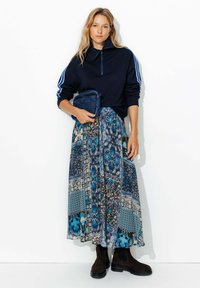 Sweat-shirt zippé bleu marine avec des rayures blanches, associé à une longue jupe à motifs aux tons bleu, marron et crème. Bottes à talons marron.