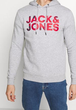 Homme portant un sweat à capuche gris clair avec le texte rouge « JACK & JONES » et un jean bleu, debout, les bras détendus le long du corps.