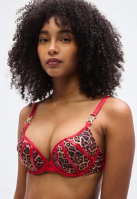 Soutien-gorge imprimé léopard avec bordure rouge et bretelles réglables. Il présente des bonnets moulés, des accents dorés et un motif géométrique sur le tissu.