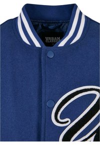 Jachetă varsity albastră cu guler cu dungi albe, închidere cu nasturi, și mare literă neagră brodată pe partea din față. Textură moale din amestec de lână.
