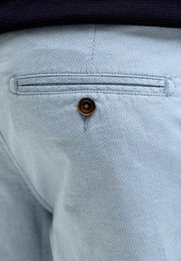 Ανοιχτό μπλε chinos με υφασμένο μοτίβο, διαθέτοντας πίσω τσέπη με καφέ κουμπί. Το ύφασμα φαίνεται μαλακό με λεπτές λεπτομέρειες.