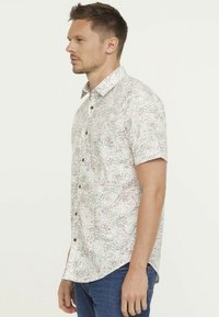 Chemise à manches courtes avec un fond crème clair, présentant un motif de feuilles vertes et brunes, et des boutons bruns sur le devant.