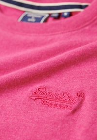 T-shirt in cotone rosa con collo rotondo, caratterizzato da un logo "Superdry" ricamato sul davanti. Il tessuto ha una consistenza liscia.