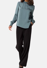 Blouse bleu sarcelle avec un col montant et des poignets froncés, associée à un pantalon large noir. Texture lisse, coupe décontractée et détails minimalistes.