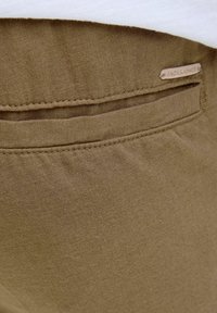 Khaaki tkanina s glatkom teksturom, sa džepom i malom oznakom "JACK & JONES" prošivenom na pojasu. Uredni detalji šivanja.