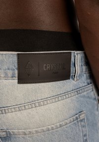 Jasnoniebieskie dżinsy z denimu z czarną skórzaną metką z napisem "CRYSTAL PARIS". Posiadają wyraźny wzór przeszyć i klasyczny pięć kieszeniowy design.