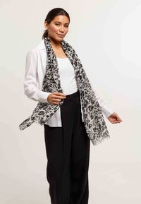 Écharpe en imprimé léopard noir et blanc en tissu l�éger, avec des bords à franges, présentée drapée autour du cou sur une chemise blanche.