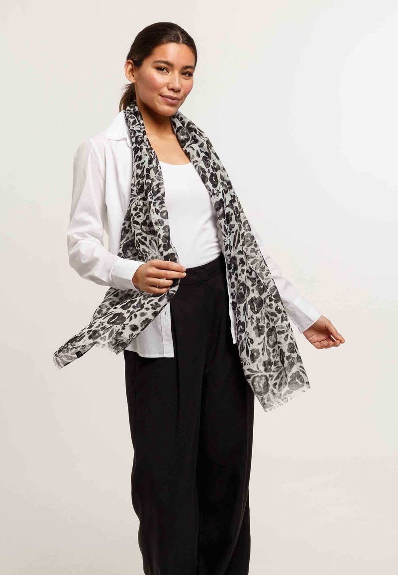 Écharpe en imprimé léopard noir et blanc en tissu léger, avec des bords à franges, présentée drapée autour du cou sur une chemise blanche.