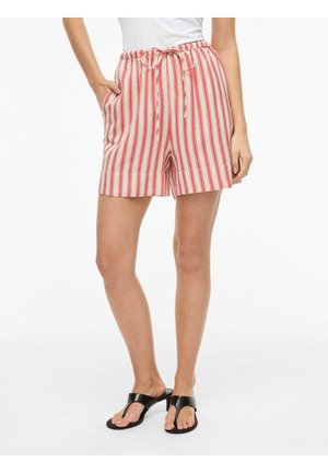 Kobieta w czerwono-białych, pionowo paskowanych szortach z troczkiem w pasie i czarnych sandałach na obcasie, stoi z jedną ręką w kieszeni.