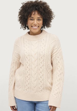 Pull en maille torsadée de couleur beige clair, présentant un motif texturé, un col et des poignets côtelés, ainsi qu'une coupe oversize. Assorti avec un jean bleu.