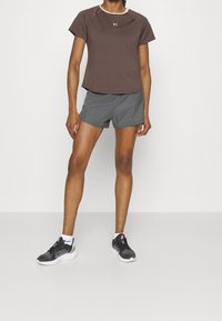 Kvinna klädd i en brun kortärmad topp, grå träningsshorts, vita strumpor och svarta löparskor står mot en neutral bakgrund.