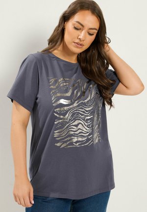 Femme aux longs cheveux bruns portant un t-shirt gris foncé avec un imprimé abstrait zèbre argenté brillant, debout sur un fond clair uni.
