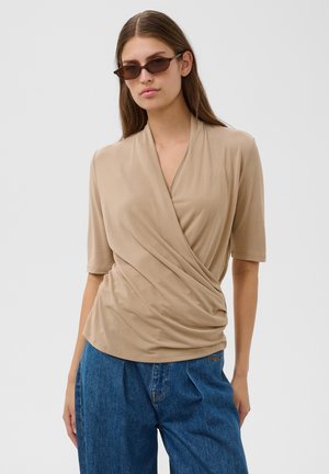 Kvinde iført beige slå-om kortærmet top, blå højtaljede jeans og mørke solbriller på en lys baggrund.