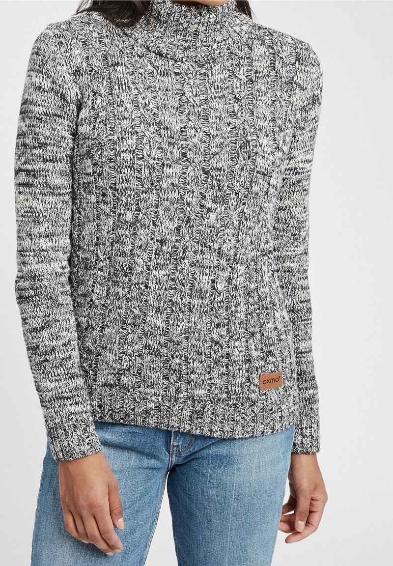 Pull à col roulé en maille noir et blanc, présentant un motif texturé et une étiquette logo en cuir marron à l'ourlet. Accompagné d'un jean bleu.