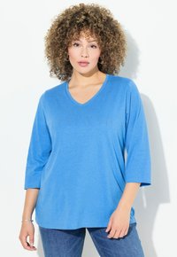 Blaues V-Ausschnitt Langarmoberteil aus weichem Stoff, mit entspannter Passform. Kombiniert mit Jeans auf einem hellen Hintergrund.