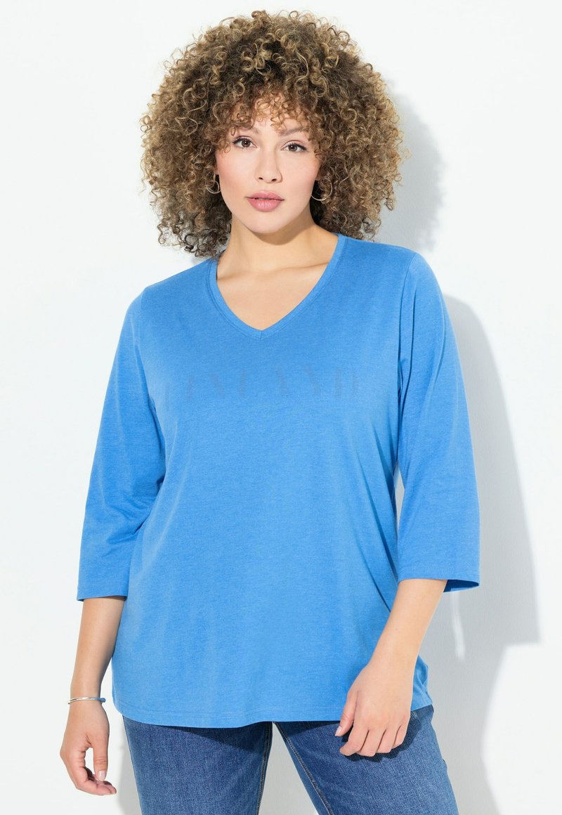 Blaues V-Ausschnitt Langarmoberteil aus weichem Stoff, mit entspannter Passform. Kombiniert mit Jeans auf einem hellen Hintergrund.