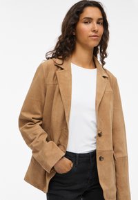 Brown suede blazer with lapel collar, two buttons, and side pockets, paired with a white top and dark jeans. Smooth texture, tailored fit.