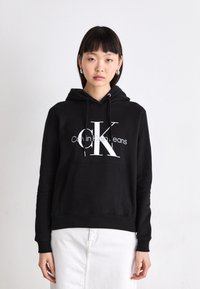 Juoda su gobtuvu pagaminta medvilnės mišinio, su dideliu baltu "CK" logotipu ir "Calvin Klein Jeans" tekstu. Ripsuoti kraštai ir rankogaliai.