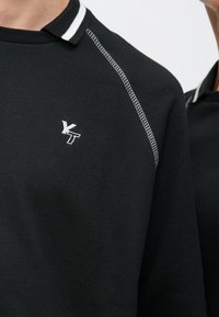 Μαύρο φούτερ με λευκή ριγέ λαιμόκοψη, αντίθετη ραφή και μικρό κεντημένο λογότυπο στο στήθος. Κοντινή λήψη.