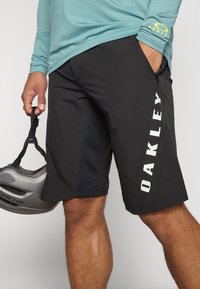 Juodos sportinės šortai su šoniniu užtrauktuku, su baltu "OAKLEY" ženklu vertikaliai kairėje kojų pusėje. Lygus audinys su prigludusia konstrukcija.