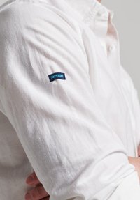 Hvid skjorte med knapper lavet af letvægtsstof, med lange ærmer og et blåt "SUPERDRY" mærke på venstre arm. Struktureret overflade.