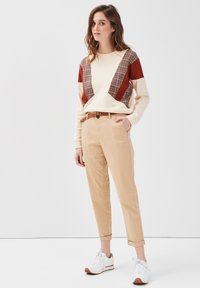 Cremefarvet sweatshirt med røde og ternede accenter, parret med beige tapered bukser og hvide sneakers med brune såler.