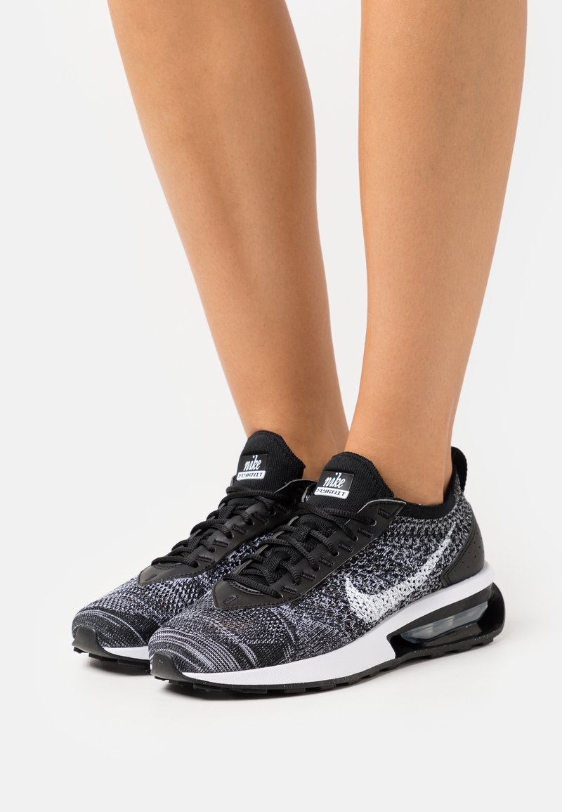 Pieds portant des baskets Nike tricotées noir et blanc avec logo swoosh blanc et amorti d'air visible dans la semelle sur un fond clair.