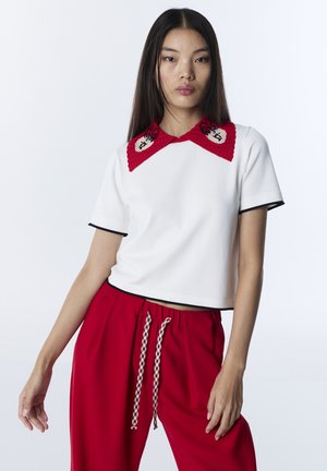 Junge Frau trägt ein weißes Kurzarmshirt mit rotem, detailliertem Kragen und rote Hose mit gemustertem Kordelzug, steht vor weißem Hintergrund.