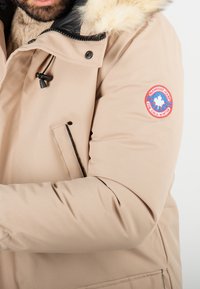 Veste doudoune beige à capuche avec des accents noirs, bordure en fourrure, un écusson sur la manche et une poche zippée à l'avant. Texture lisse.