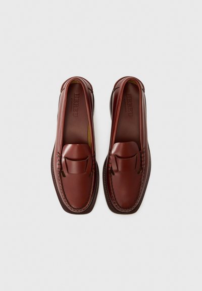 Braune Lederloafers mit einem Mokassin-Design, ausgestattet mit einem gesteppten Oberteil, flacher Sohle und einem dekorativen Riemen über der Vorderseite.